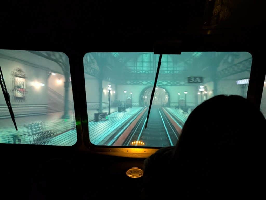 Blick aus dem Führerstand einer Eisenbahnsimulation auf einen nebligen, unterirdischen Bahnhof mit beleuchteten Bahnsteigen und Gleisen.