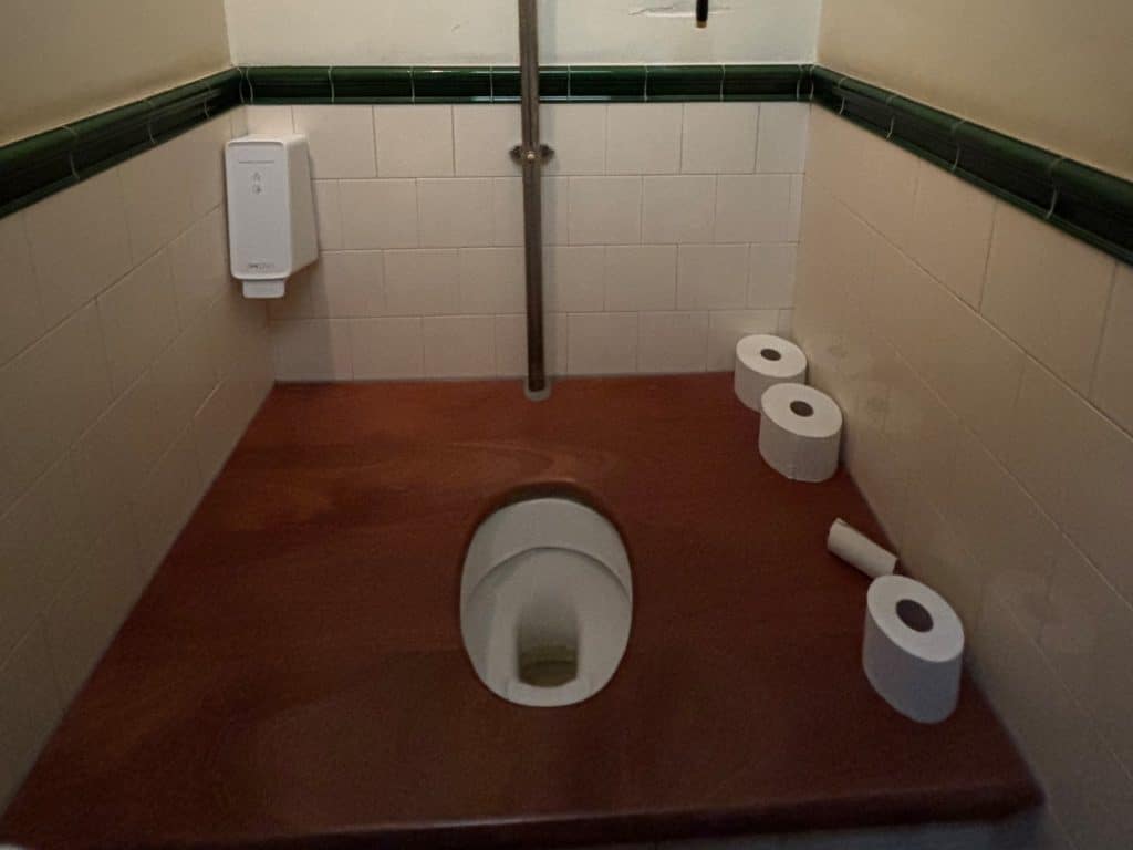 Eine Toilette mit einer großen, rötlichen Holzbank, die eine zentrale Öffnung über dem Toilettenbecken besitzt. Die Wände sind hell gefliest und mit einer grünen Zierleiste versehen, während mehrere Rollen Toilettenpapier auf der Holzfläche bereitliegen.