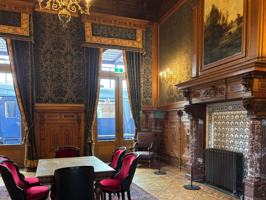 Ein prunkvoller historischer Wartesaal im Eisenbahnmuseum Utrecht mit holzgetäfelten Wänden, gemusterten Tapeten, einem kunstvollen Kamin und einer Sitzgruppe aus roten Samtstühlen vor einem Fenster mit Blick auf einen Zug.