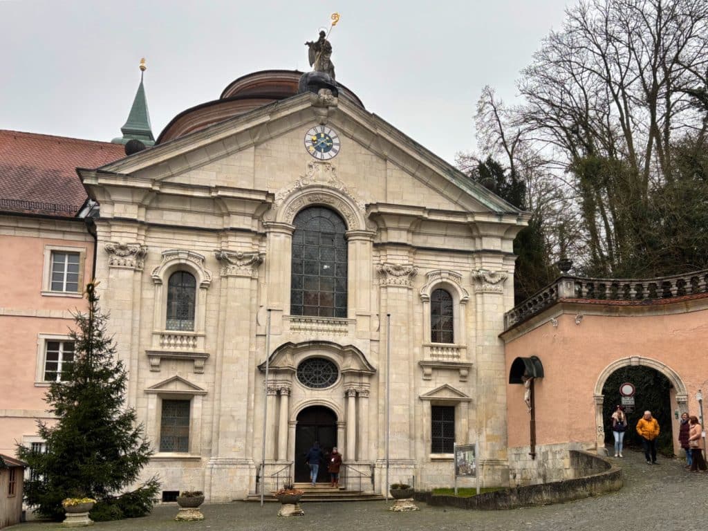 Die barocke Steinfassade der Klosterkirche Weltenburg mit einer goldenen Statue auf dem Giebel, flankiert von einem Weihnachtsbaum und einem Torbogen mit Passanten.