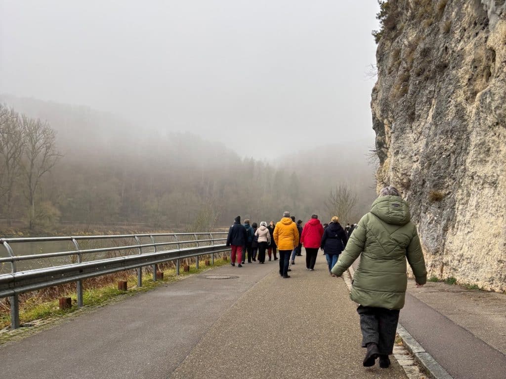 Eine Gruppe von Spaziergängern läuft an einem nebligen Tag auf dem Weltenburger Fußweg entlang der Donau, gesäumt von einer hohen Felswand und herbstlichen Bäumen im Hintergrund.