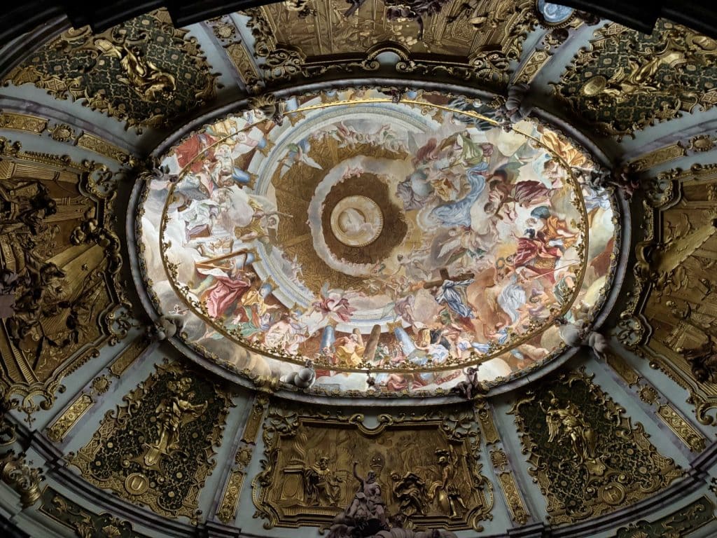 Blick nach oben in die prachtvolle Deckenkuppel der Klosterkirche Weltenburg, die ein farbenfrohes Fresko mit zahlreichen Figuren umgeben von goldenen Stuckverzierungen und Reliefs zeigt.