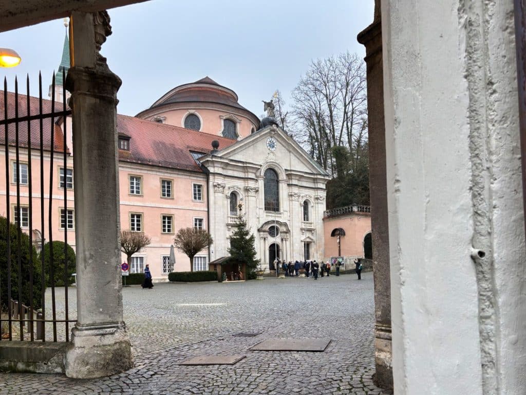 Blick durch einen steinernen Torbogen auf den gepflasterten Innenhof des Klosters Weltenburg mit seiner barocken Klosterkirche und den angrenzenden rosafarbenen Gebäuden unter bewölktem Himmel.