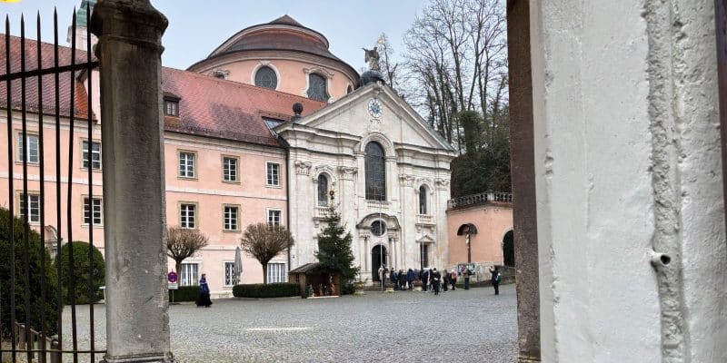 Blick durch einen steinernen Torbogen auf den gepflasterten Innenhof des Klosters Weltenburg mit seiner barocken Klosterkirche und den angrenzenden rosafarbenen Gebäuden unter bewölktem Himmel.