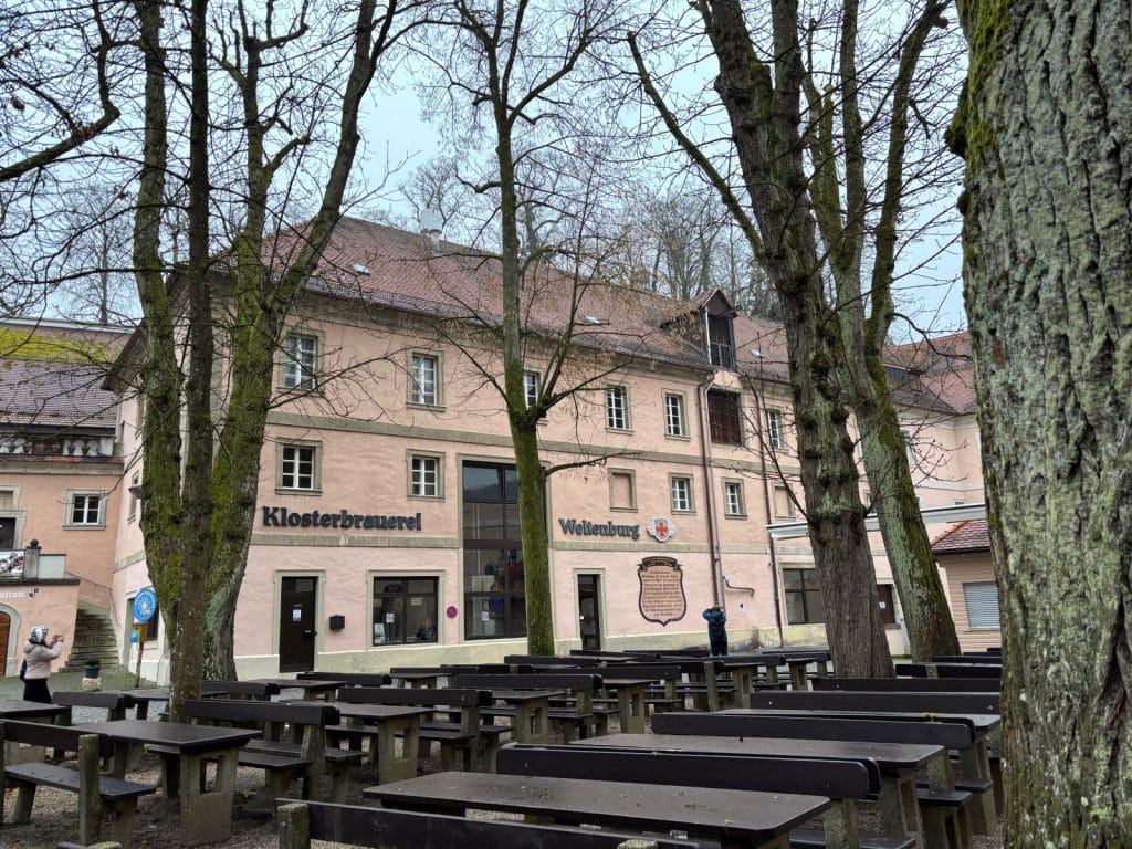 Das Bild zeigt das mehrstöckige, rosafarbene Gebäude der Klosterbrauerei Weltenburg mit einem vorgelagerten Biergarten voller leerer Holztische und Bänke unter hohen, kahlen Bäumen. An der Fassade sind die Schriftzüge „Klosterbrauerei“ und „Weltenburg“ sowie ein historisches Wappen und eine Informationstafel angebracht.