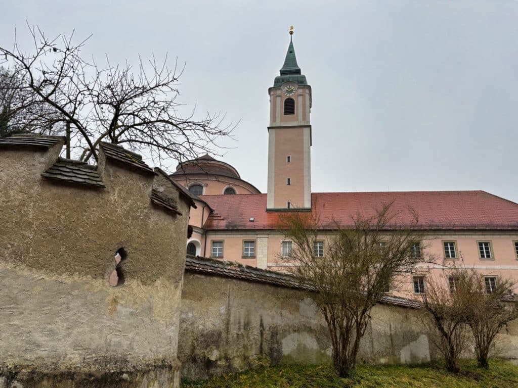 Blick über eine historische Steinmauer auf die barocke Klosteranlage Weltenburg mit ihrer rosafarbenen Fassade, dem markanten Kirchturm und der Kuppel unter grauem Himmel.