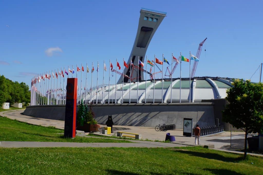 Olympische Spiele Montreal: Das Montrealer Olympiastadion mit seinem markanten schrägen Turm und einer Reihe wehender internationaler Flaggen vor einem klaren blauen Himmel. Im Vordergrund erstreckt sich eine Grünfläche mit einem gepflasterten Weg, auf dem ein Fahrradständer und eine Person zu sehen sind.