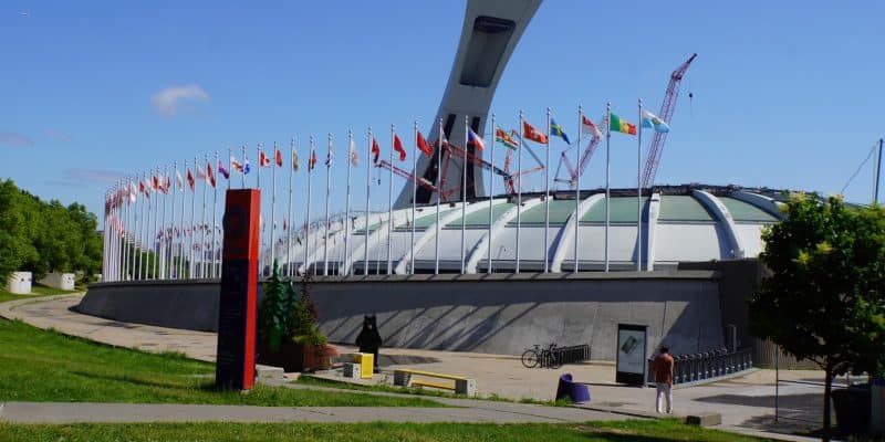 Das Montrealer Olympiastadion mit seinem markanten schrägen Turm und einer Reihe wehender internationaler Flaggen vor einem klaren blauen Himmel. Olympische Spiele Montreal