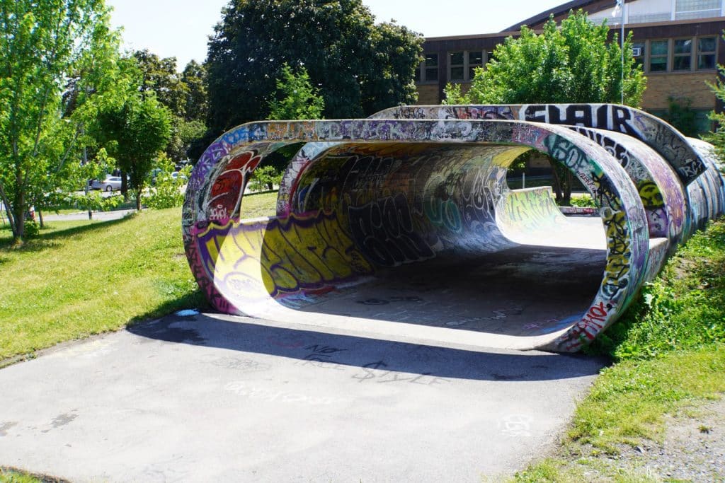 Eine mit buntem Graffiti besprühte Betonröhre im Olympiapark von Montreal liegt auf einer asphaltierten Fläche inmitten einer grünen Parkanlage. Die röhrenförmige Skulptur ist an den Seiten offen und gibt den Blick auf weitere dahinterliegende Betonelemente und Bäume frei.