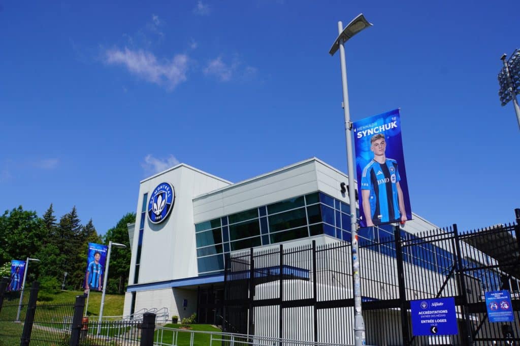 Außenansicht des Montrealer Olympiastadions bei blauem Himmel, mit dem Vereinslogo des CF Montréal an der weißen Fassade und blauen Werbebannern von Fußballspielern an den Laternenmasten.