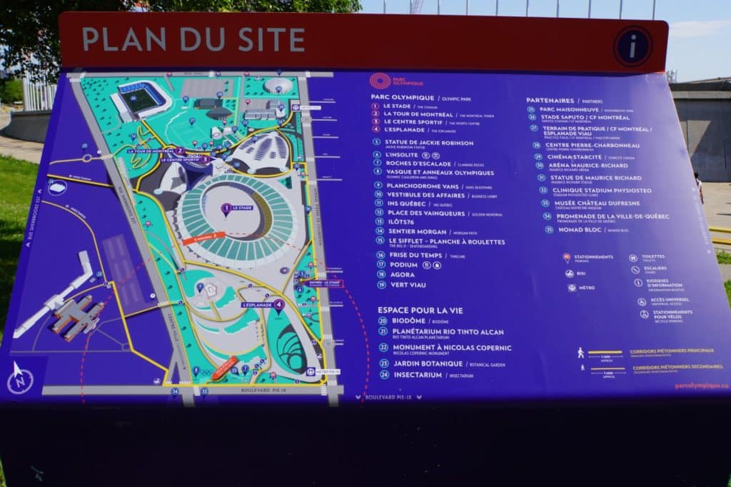 Ein blau-roter Plan des Montrealer Olympiaparks zeigt eine detaillierte Karte des Geländes mit dem Olympiastadion im Zentrum sowie eine nummerierte Legende der Sehenswürdigkeiten und Partnerstandorte. Der Plan enthält zudem Symbole für Parkplätze, Toiletten und Metrostationen sowie Informationen zum Espace pour la vie, wie dem Biodôme und dem Botanischen Garten.