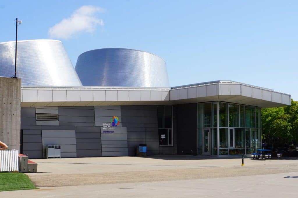 Das Rio Tinto Alcan Planetarium in Montreal zeigt zwei markante, silberne kegelförmige Türme über einem modernen, flachen Gebäude mit großen Glasfronten unter blauem Himmel.