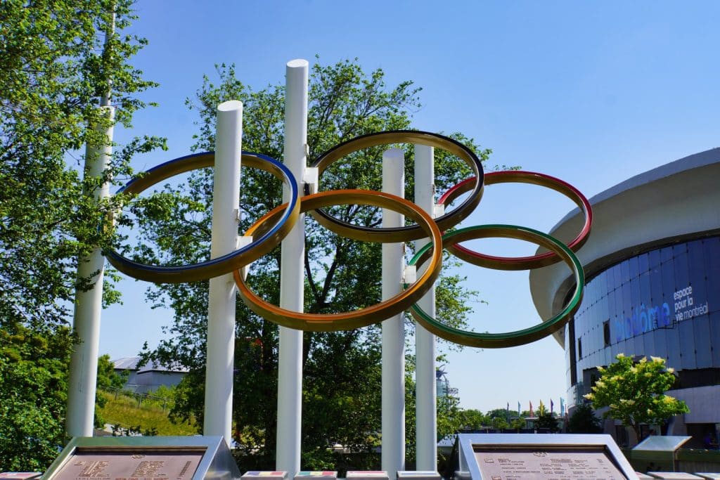 Olympische Spiele Montreal: Eine Skulptur der fünf olympischen Ringe steht auf weißen Säulen vor dem Biodôme in Montreal unter einem klaren blauen Himmel. Im Vordergrund befinden sich Informationstafeln, während im Hintergrund grüne Bäume und ein Teil des modernen Gebäudes zu sehen sind.