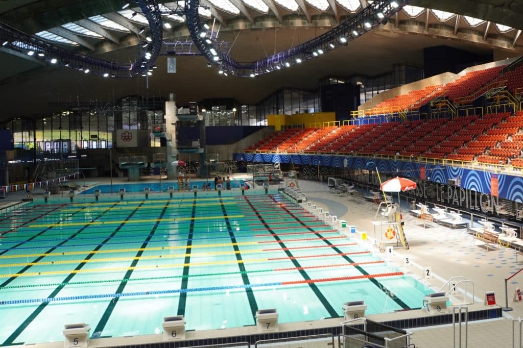 Olympische Spiele Montreal: Innenansicht der Montreal Olympia Schwimmhalle mit einem großen Wettkampfbecken, Startblöcken, Sprungtürmen im Hintergrund und leeren, orangefarbenen Zuschauerrängen unter einer markanten Deckenkonstruktion.