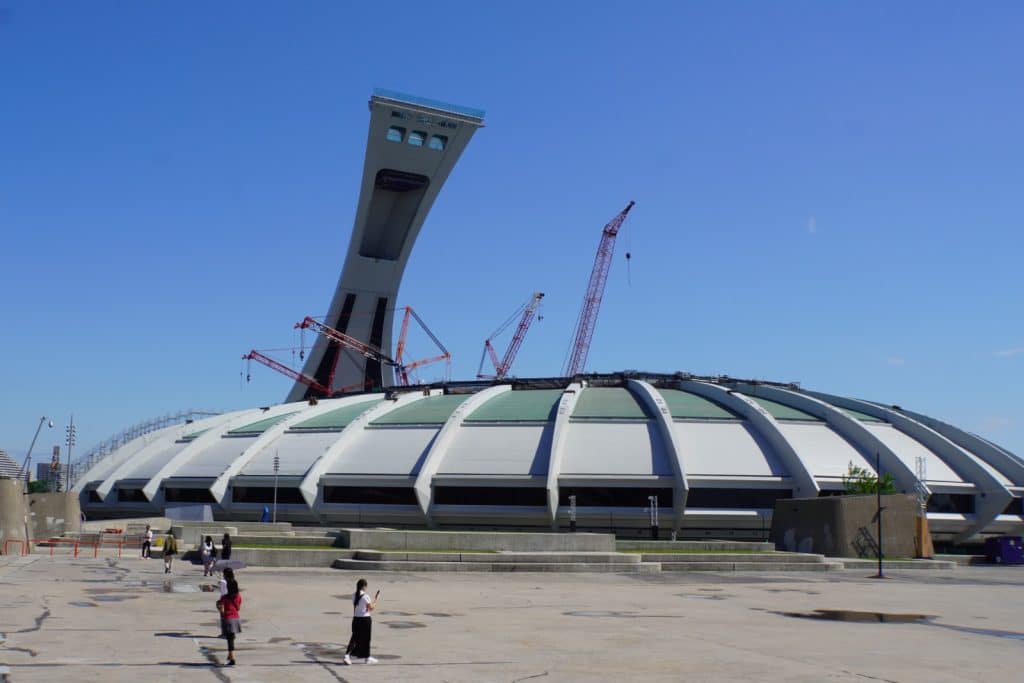Außenansicht des Olympiastadions in Montreal mit seinem markanten, geneigten Turm und der kuppelförmigen Dachstruktur, hinter der mehrere Baukräne aufragen.