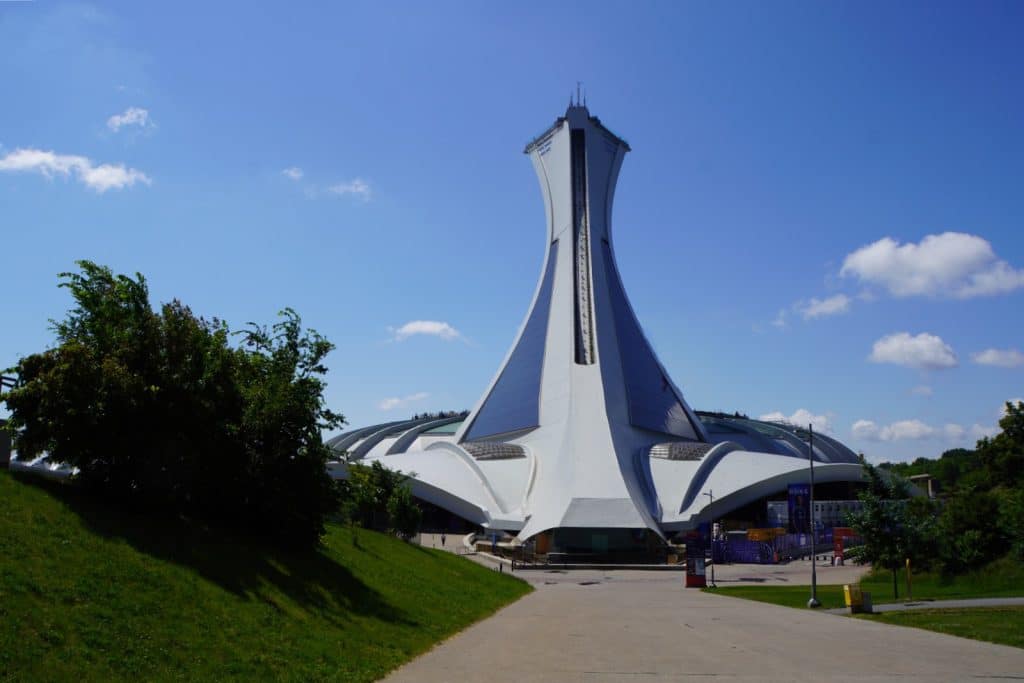 Olympische Spiele Montreal: Der weiße Montrealer Olympiaturm ragt mit seiner markanten, geneigten Architektur hinter dem Stadiondach unter einem strahlend blauen Himmel empor. Ein gepflasterter Weg im Vordergrund führt an grünen Wiesen und Bäumen vorbei direkt auf das monumentale Bauwerk zu.