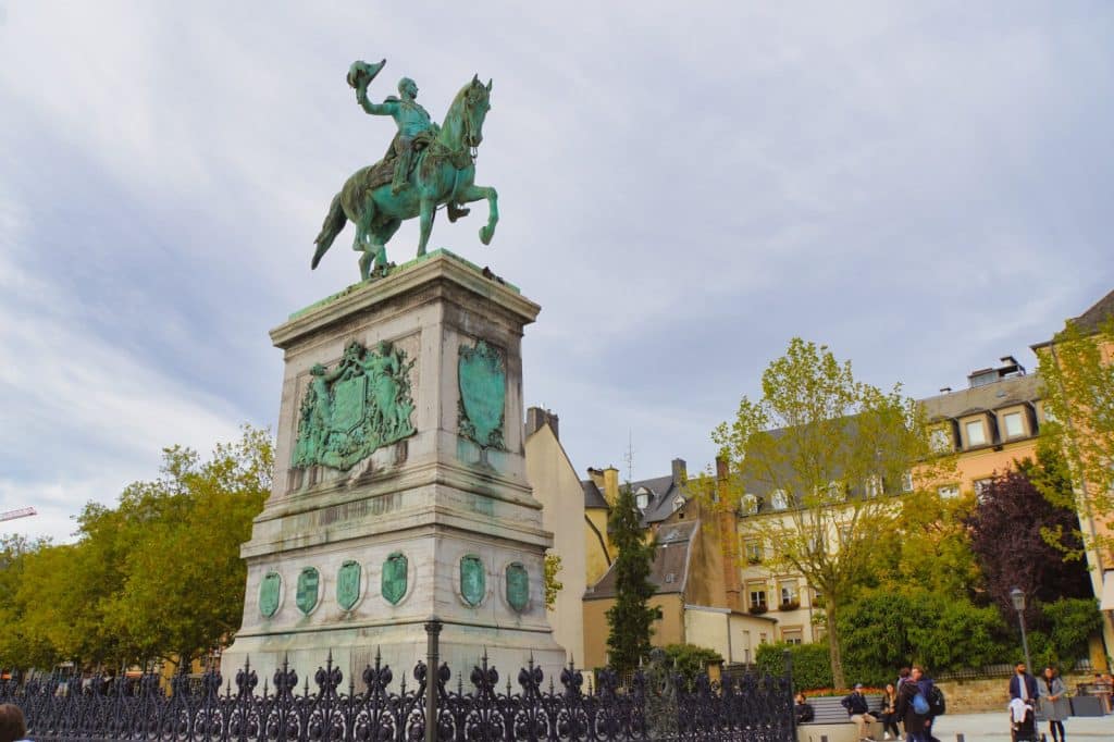 Das bronzene Reiterstandbild von König-Großherzog Wilhelm II. thront auf einem hohen Steinsockel mit Wappenreliefs auf dem Place Guillaume II in Luxemburg-Stadt, umgeben von Bäumen und historischen Gebäuden.
