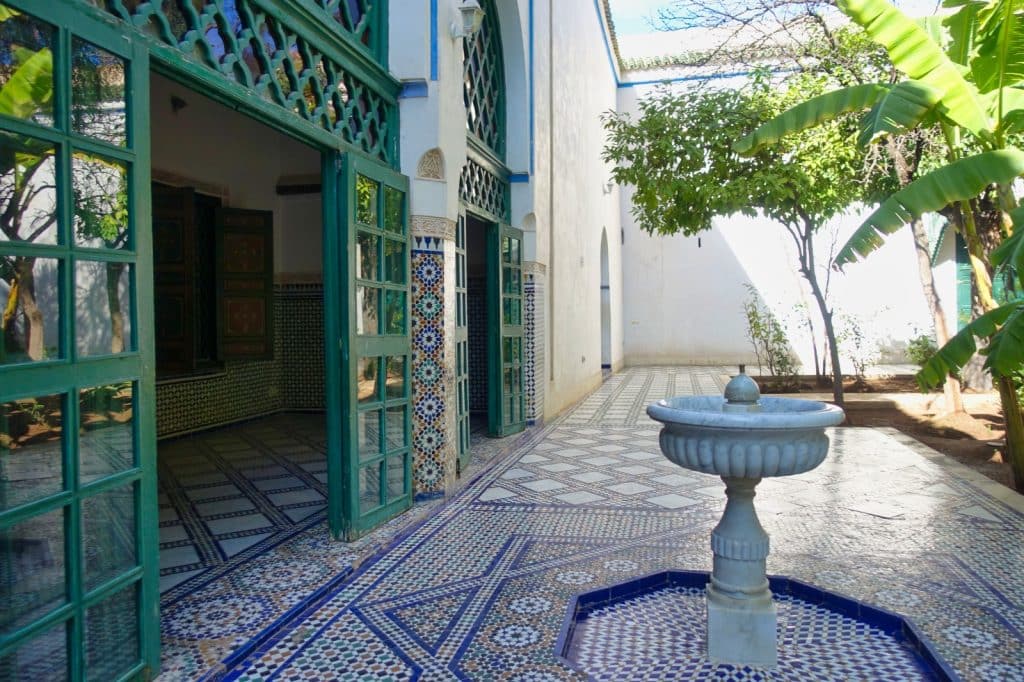 Ein sonniger Innenhof im Großen Riad des Bahia-Palasts in Marrakesch mit kunstvollen Mosaikböden, einer zentralen Marmorfontäne und grünen Holztüren, die in schattige Räume führen.