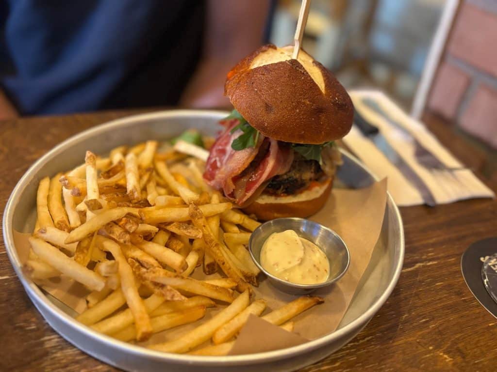 Ein Burger der Brauerei Les 3 Brasseurs in Montreal mit Speck und Rucola auf einem Laugenbrötchen, serviert mit einer großen Portion Pommes frites und einem Dip in einer Metallschale.