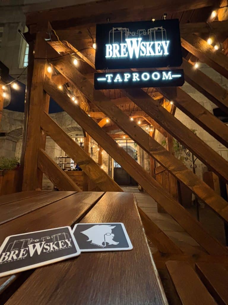 Abendliche Außenansicht des Brewskey Taproom in Montreal mit beleuchteten Schildern über einem hölzernen Eingangsbereich und zwei passenden Untersetzern auf einem rustikalen Holztisch im Vordergrund.
