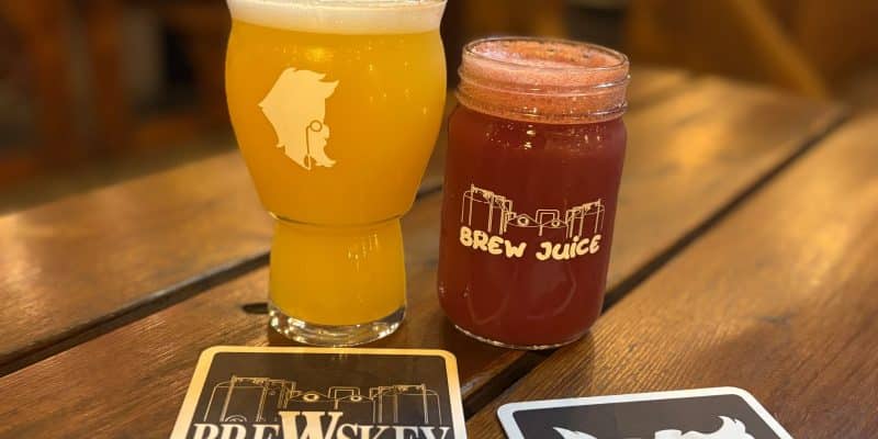 Zwei Gläser mit Brauerei im Vieux Port in Montreal stehen auf einem Holztisch neben passenden Untersetzern, wobei ein Glas ein hellgelbes, trübes Bier und das andere ein rötliches „Brew Juice“ enthält.