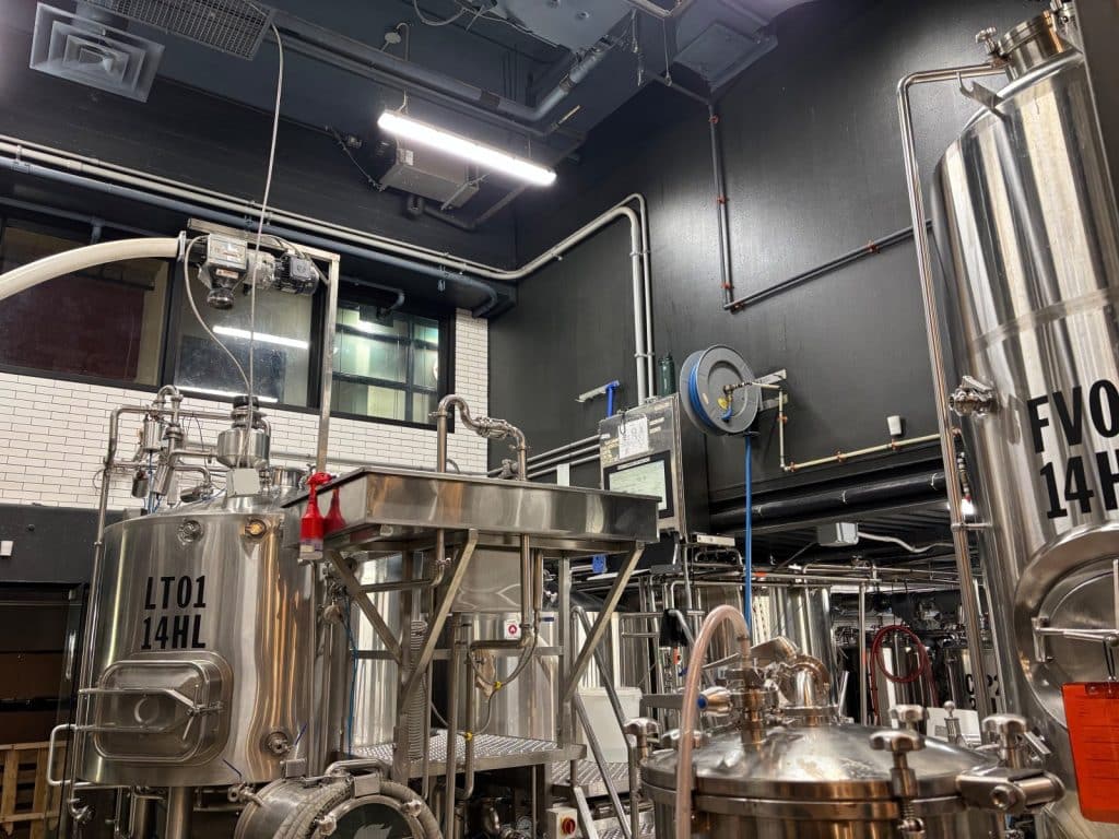 Industrielle Braukessel aus glänzendem Edelstahl mit Beschriftungen wie „LT01 14HL“ stehen in der modernen Produktionshalle der Montreal Brewskey Brauerei unter einer dunklen Decke mit sichtbaren Rohrleitungen.
