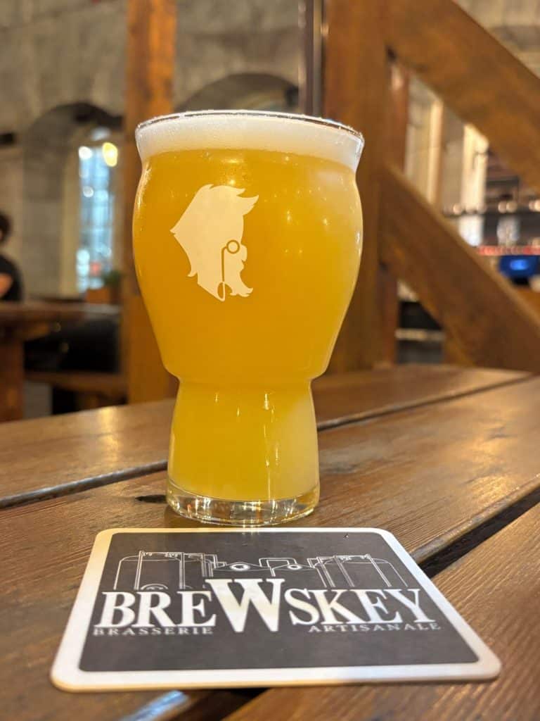 Ein Glas trübes, gelbes Bier mit weißem Schaum steht auf einem Holztisch der Brauerei im Vieux Port in Montreal, direkt neben einem passenden Bierdeckel mit dem Logo der Brauerei.