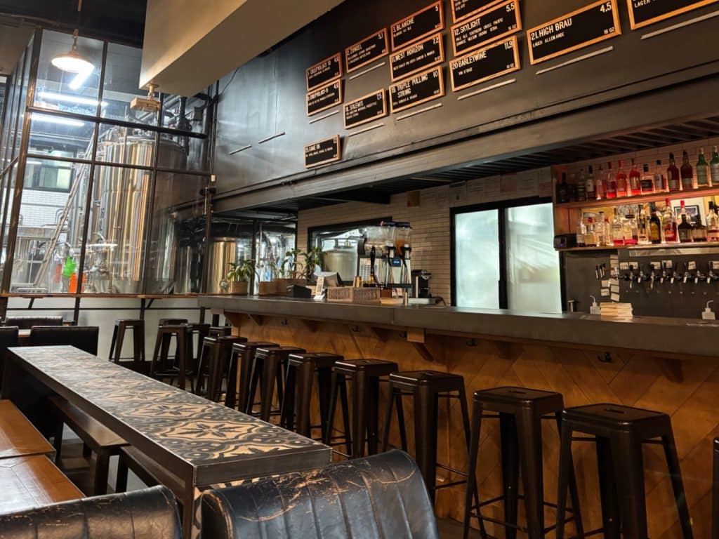 Innenansicht der Brauerei Brewskey in Montreal mit einer langen Bar aus Holz, hohen Metallhockern und einer großen Auswahl an Getränken auf Wandtafeln, während im Hintergrund glänzende Braukessel durch eine Glaswand sichtbar sind.
