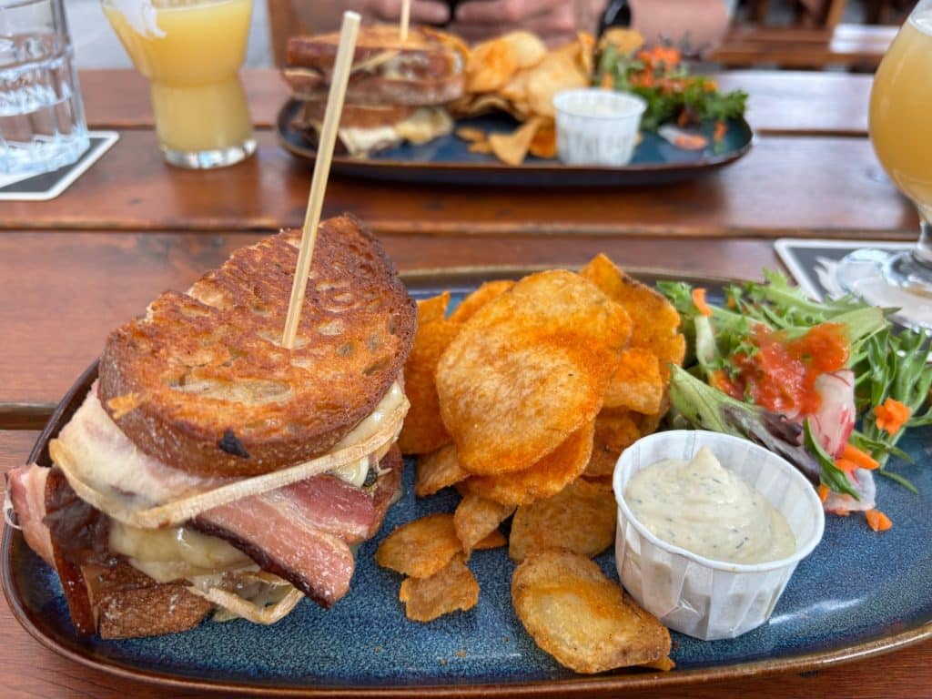 Ein reichhaltig belegtes Sandwich mit Speck und Käse von Brewskey in Montreal, serviert auf einem blauen Teller mit knusprigen Kartoffelchips, einem kleinen Beilagensalat und einem Dip. Im Hintergrund ist ein zweiter Teller mit einem ähnlichen Gericht sowie Gläser mit Bier auf einem Holztisch zu sehen.