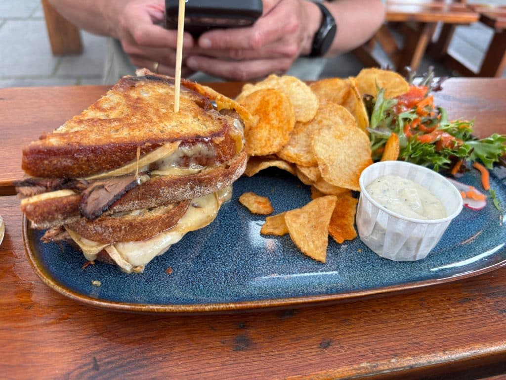 Ein Sandwich mit geschmolzenem Käse und Fleisch auf geröstetem Brot, serviert auf einem blauen Teller mit Kartoffelchips, einem Beilagensalat und einem kleinen Becher Dip.