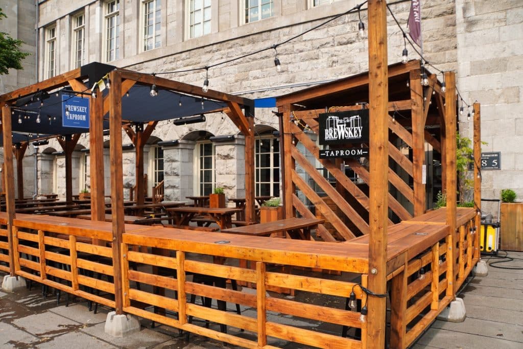 Außenansicht der hölzernen Terrasse des Brewskey Taproom in Montreal mit rustikalen Holzbänken, Tischen und Lichterketten vor einem historischen Steingebäude.