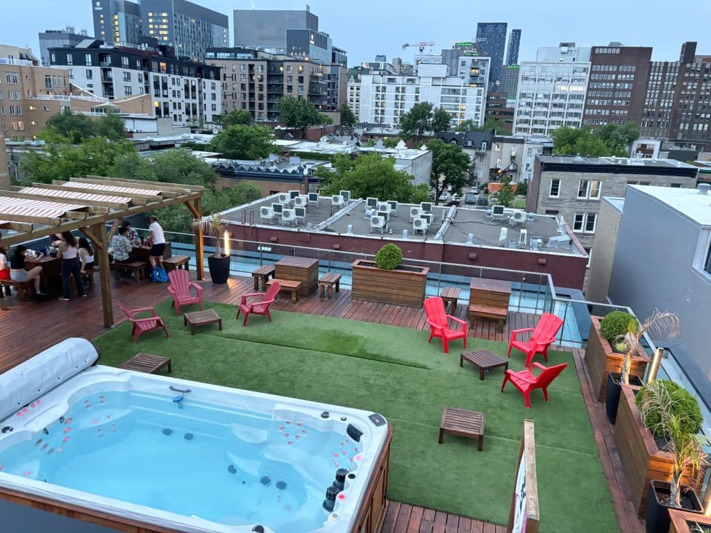 Dachterrasse coolste Hostel in Montréal mit einem großen Whirlpool im Vordergrund, einer Kunstrasenfläche mit roten Sesseln und einer Bar unter einer Pergola vor der städtischen Skyline.