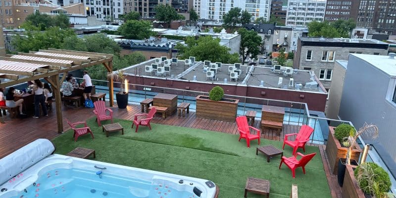 Dachterrasse coolste Hostel in Montréal mit einem großen Whirlpool im Vordergrund, einer Kunstrasenfläche mit roten Sesseln und einer Bar unter einer Pergola vor der städtischen Skyline.
