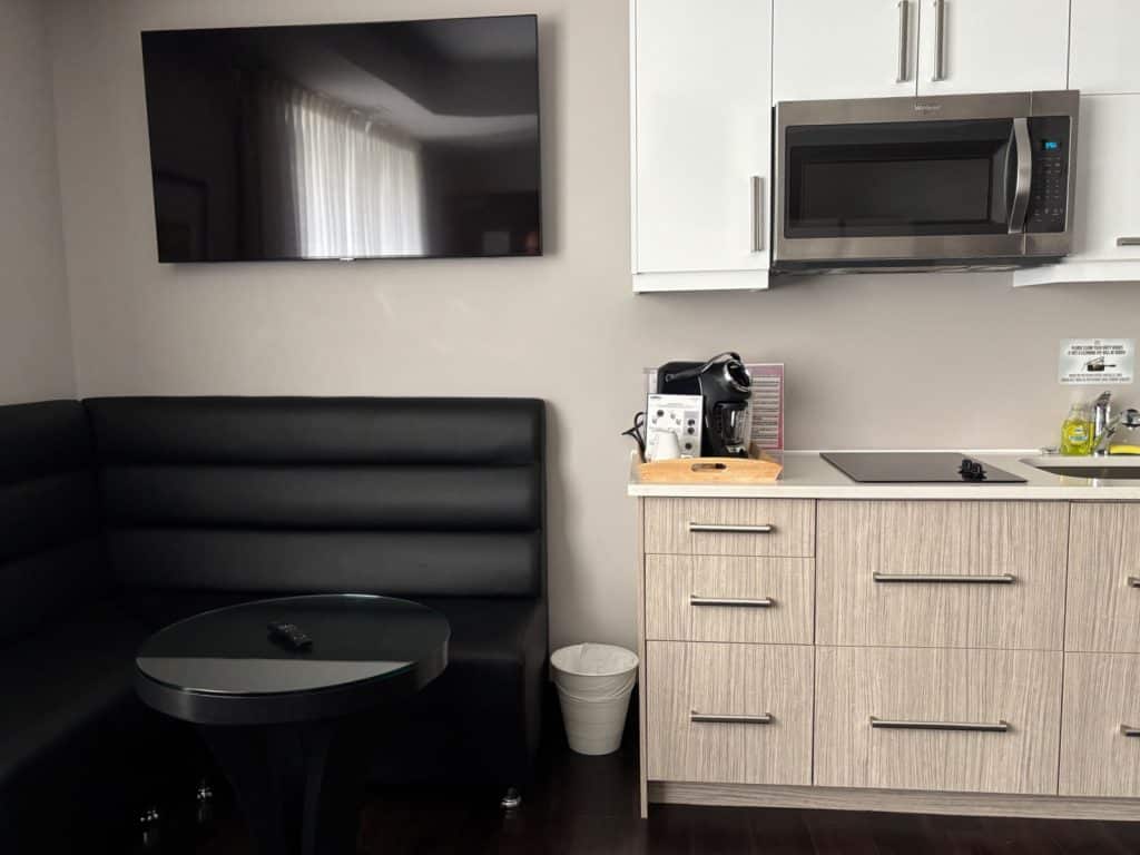 Eine moderne Küchenzeile im M Montreal mit hellen Holzschränken, einer Mikrowelle aus Edelstahl und einer schwarzen Ledersitzecke mit passendem runden Tisch. Über der Sitzbank ist ein großer Flachbildfernseher an der Wand montiert.