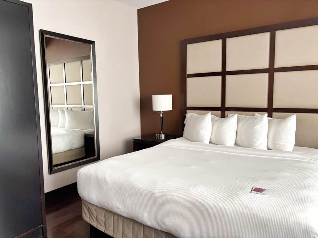 Ein Hostelzimmer im M Montreal mit einem großen Bett mit weißer Bettwäsche, einem gepolsterten Kopfteil in Gitteroptik vor einer braunen Akzentwand und einem großen Wandspiegel.