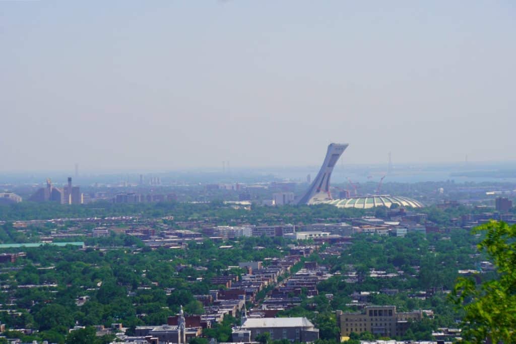 Weitläufiger Blick vom Mont Royal über Montreal auf das markante Olympiastadion mit seinem geneigten Turm, eingebettet in ein dichtes Stadtviertel mit viel Grün.