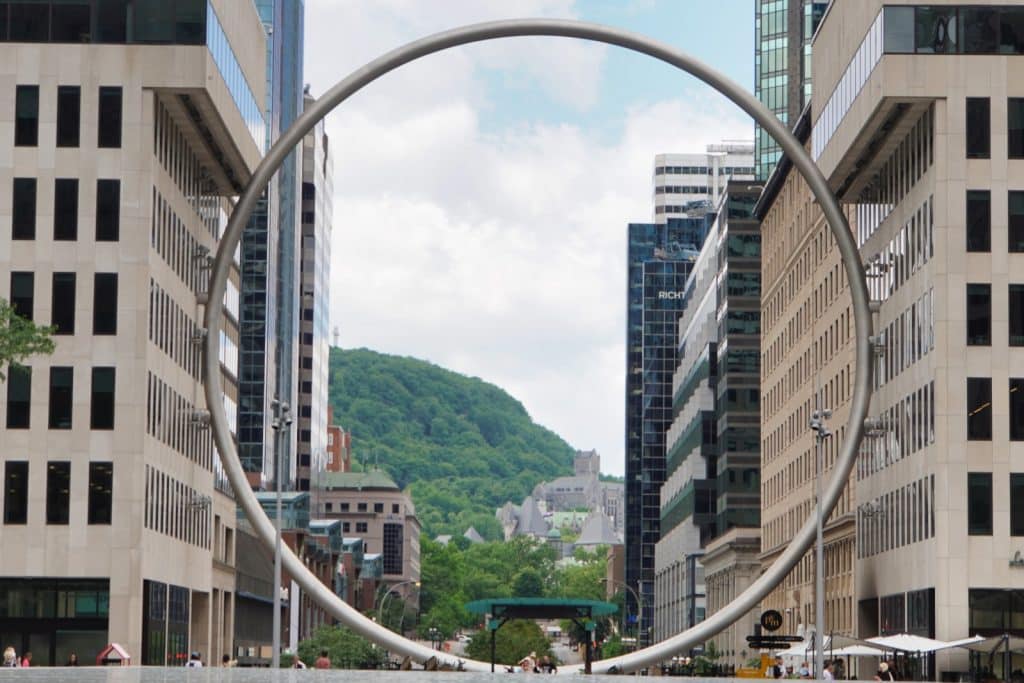 Eine große, kreisförmige Metallskulptur rahmt den Blick durch eine Häuserschlucht in Montreal auf den bewaldeten Mont-Royal und die umliegende Stadtarchitektur ein.