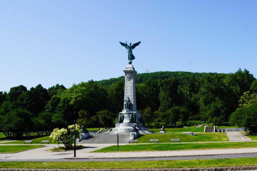 Das George-Étienne-Cartier-Monument am Fuße des Mont Royal in Montreal zeigt eine hohe Steinsäule mit einer geflügelten Bronzestatue an der Spitze, umgeben von einer Parklandschaft mit Bäumen und Gehwegen.