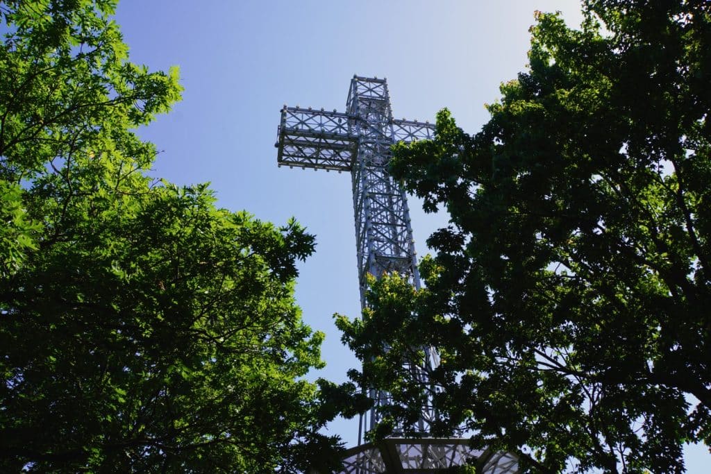 Ein hohes, aus einer Stahlgitterkonstruktion gefertigtes Kreuz ragt auf dem Mont Royal in Montreal zwischen dichten, grünen Baumkronen in den klaren blauen Himmel.