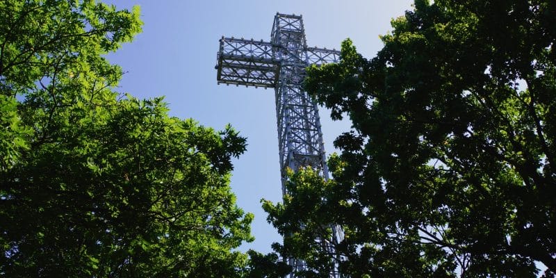 Ein hohes, aus einer Stahlgitterkonstruktion gefertigtes Kreuz ragt auf dem Mont Royal in Montreal zwischen dichten, grünen Baumkronen in den klaren blauen Himmel.