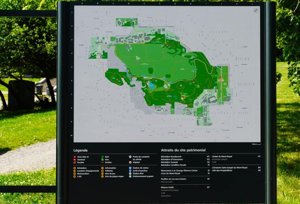 Ein großer, schwarz gerahmter Parkplan des Mont-Royal in Montreal steht im Freien vor einer grünen Parklandschaft und zeigt detailliert Wanderwege, Sehenswürdigkeiten sowie eine Legende in französischer Sprache.
