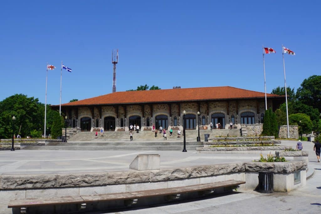 Außenansicht des Pavillons am Mont Royal in Montreal mit seiner markanten Steinfassade, dem roten Ziegeldach und einer weiten Steinterrasse, auf der Menschen spazieren gehen.