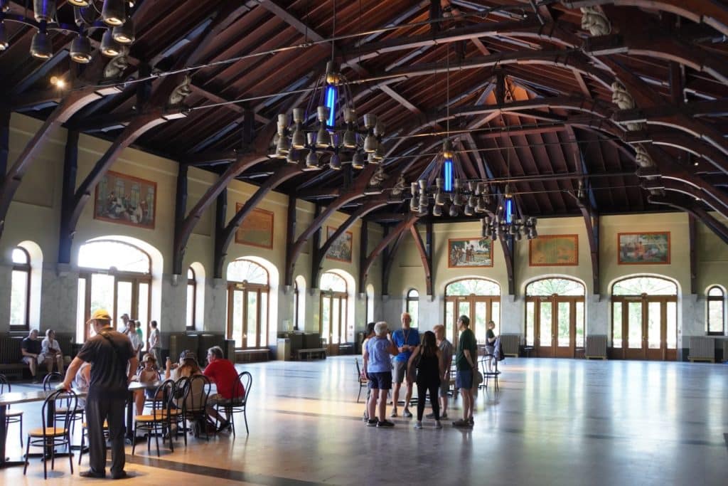 Das weitläufige Innere des Pavillons auf dem Mont Royal in Montreal besticht durch eine hohe, dunkle Holzgebälkdecke mit kunstvollen Kronleuchtern und großen Rundbogenfenstern, während sich mehrere Personengruppen im Saal aufhalten.
