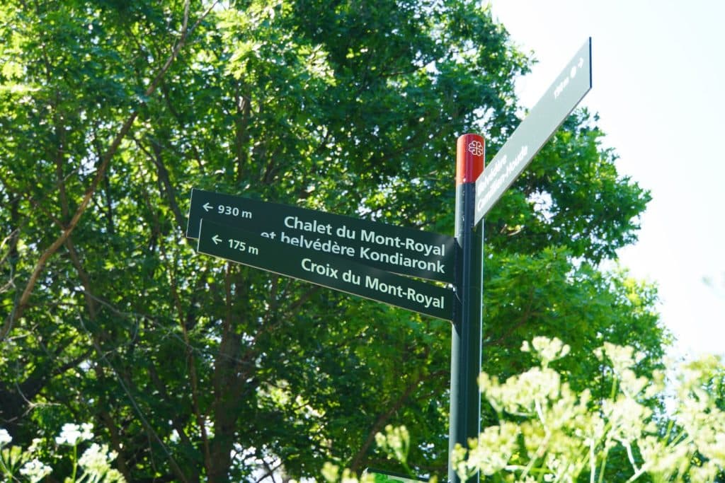 Ein grüner Wegweiser auf dem Mont Royal in Montreal zeigt Richtungen und Entfernungen zum Chalet du Mont-Royal, zum Kondiaronk-Belvedere und zum Gipfelkreuz vor einem Hintergrund aus grünen Bäumen an.