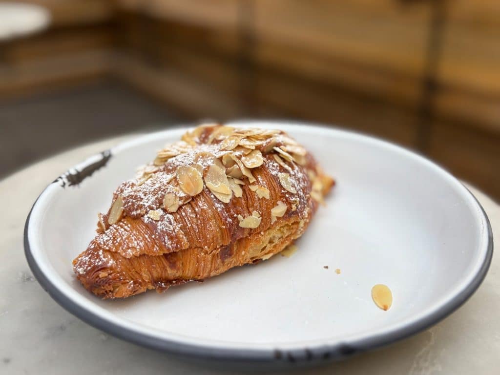 Restauranttipp Montreal: Ein frisch gebackenes, goldbraunes Mandelcroissant aus einer Bäckerei in Montreal, bestäubt mit Puderzucker und gehobelten Mandeln, serviert auf einem weißen Teller mit dunklem Rand.