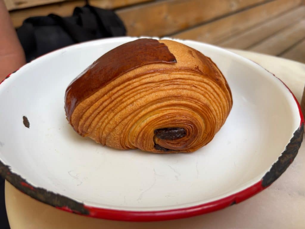 Ein frisch gebackenes Schokoladencroissant mit goldbraun glänzender Kruste und deutlich sichtbaren Teigschichten liegt in einer rustikalen weißen Emaille-Schale mit rotem Rand in einer Bäckerei in Montreal.