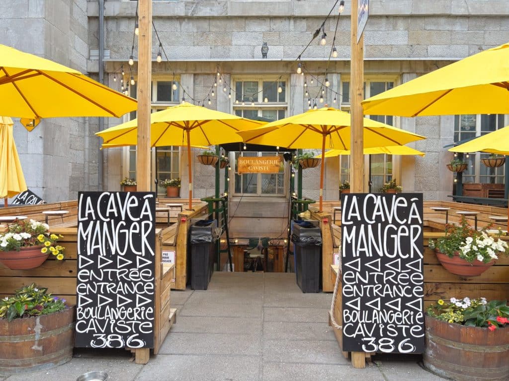 Der Eingang zur Bäckerei „La Cave à Manger“ in Montreal wird von zwei schwarzen Kreidetafeln mit weißer Aufschrift und gelben Sonnenschirmen über hölzernen Sitzbereichen flankiert. Eine Treppe führt hinunter zum Restaurant, das mit Lichterketten und Blumenkästen dekoriert ist.