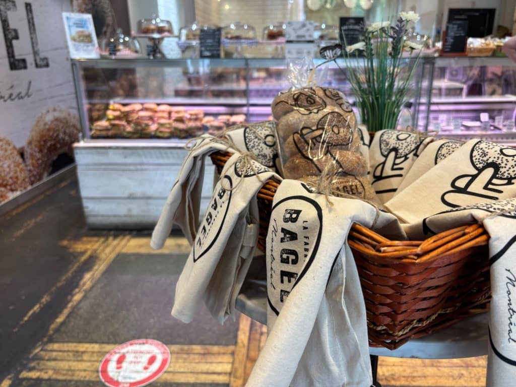 Restauranttipp Montreal: Ein Weidenkorb mit Stoffbeuteln der „La Fabrique du Bagel Montreal“ steht im Vordergrund einer Bäckerei, während im Hintergrund eine Vitrine mit Gebäck und Bagels zu sehen ist.