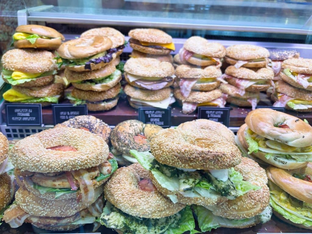 Eine Auslage in einem Restaurant in Montreal zeigt eine Vielzahl von frisch belegten Bagels, die mit Zutaten wie Omelett, Speck, Avocado und Salat gefüllt und in hohen Stapeln hinter einer Glasscheibe angerichtet sind.