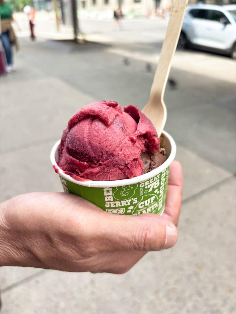 Eine Hand hält einen grünen Ben & Jerry's Pappbecher mit einer Kugel dunklem Beeren-Sorbet und einer Kugel Schokoladeneis, serviert mit einem Holzlöffel in einer Straße in Montreal.